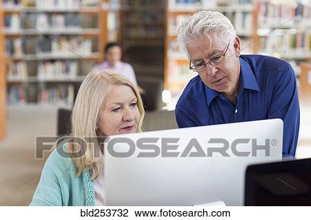 Stock Image - Older man helping woman using computer in library. Fotosearch