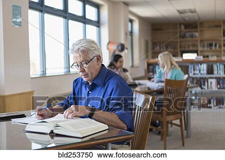 Stock Image - Older man reading book in library and writing notes. Fotosearch