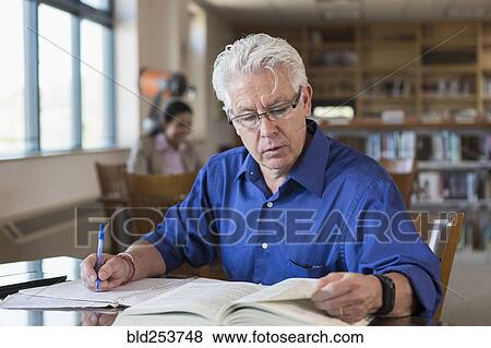 Stock Photo - Older man reading book in library and writing notes. Fotosearch