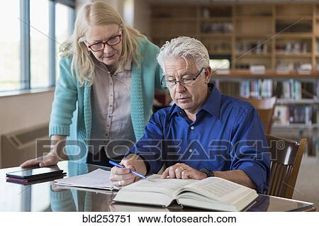 Stock Image - Older woman helping man reading book in library. Fotosearch