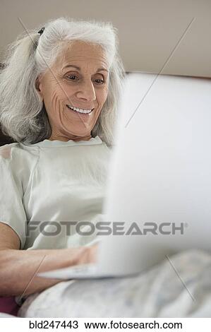 Stock Image - Older woman laying in the bed using laptop. Fotosearch
