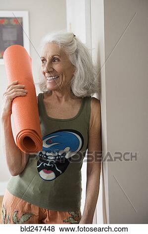 Stock Photo - Older woman leaning on wall holding exercise mat. Fotosearch