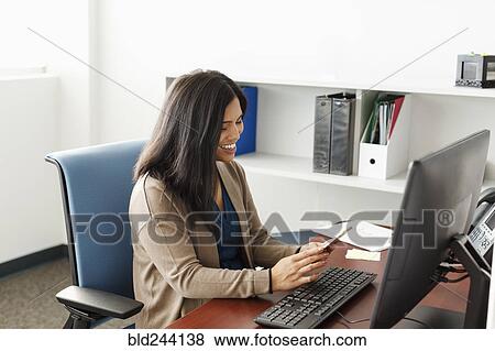 Stock Photo - Pacific Islander texting on cell phone near computer in office. Fotosearch