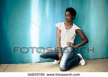 Stock Photography - Pensive African American woman kneeling near blue wall. Fotosearch