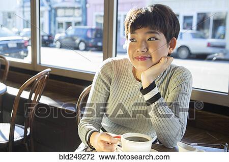 Stock Image - Pensive Asian teenage girl drinking coffee in coffee shop. Fotosearch