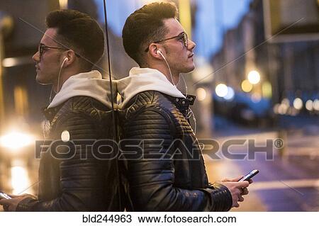 Stock Image - Pensive Caucasian man leaning on wall listening to music on cell phone. Fotosearch