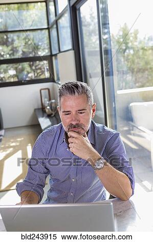 Pensive Caucasian man sitting at table using laptop View Large Photo Image Stock Photography - Pensive Caucasian man sitting at table using laptop. Fotosearch
