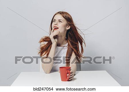 Picture - Pensive Caucasian woman sitting at windy table holding red cup. Fotosearch