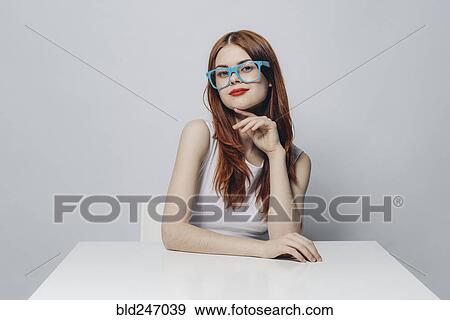 Stock Photo - Pensive Caucasian woman sitting at table wearing blue eyeglasses. Fotosearch