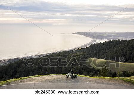 Stock Image - Person with motorcycle at edge of cliff near ocean. Fotosearch