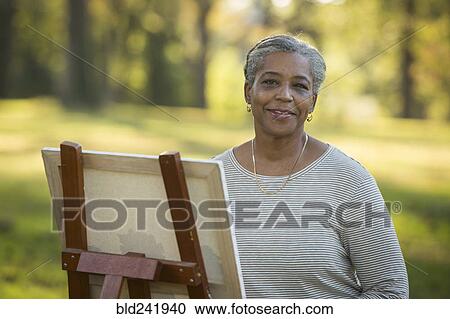 Stock Image - Portrait of Black woman painting on canvas in park. Fotosearch