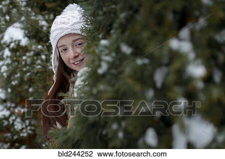 Stock Image - Portrait of Caucasian girl hiding behind tree in winter. Fotosearch
