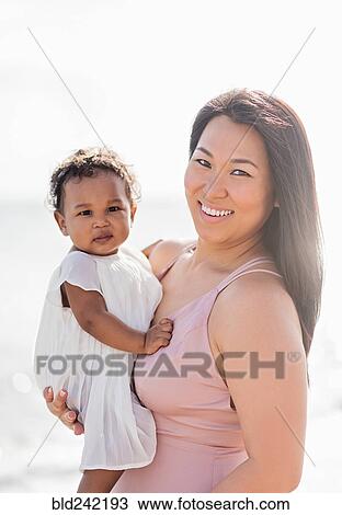 Stock Image - Portrait of mother holding baby daughter near ocean. Fotosearch