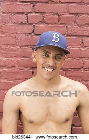 Stock Image - Portrait of shirtless Mixed Race man wearing hat near brick wall. Fotosearch