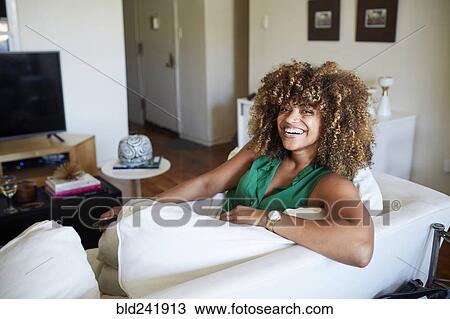 Stock Image - Portrait of smiling Black woman sitting on sofa. Fotosearch