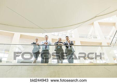 Stock Image - Portrait of smiling business people leaning on railing in lobby. Fotosearch
