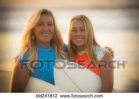 Stock Image - Portrait of smiling Caucasian mother and daughter holding surfboards. Fotosearch