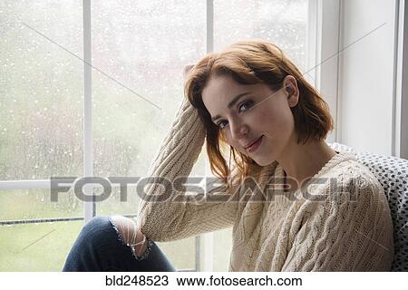 Stock Image - Portrait of smiling Caucasian woman near rainy window. Fotosearch