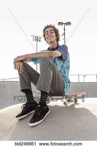 Stock Image - Portrait of smiling Hispanic man sitting on skateboard. Fotosearch