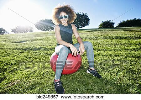 Stock Photo - Portrait of smiling Hispanic woman sitting on fitness ball in field. Fotosearch