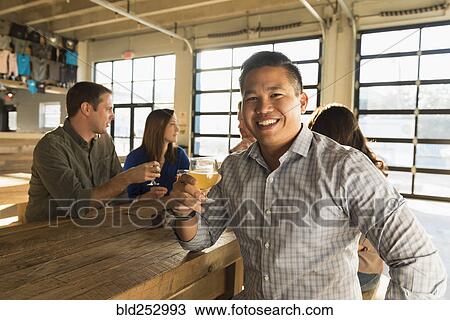 Stock Image - Portrait of smiling man drinking beer with friends in brew pub. Fotosearch