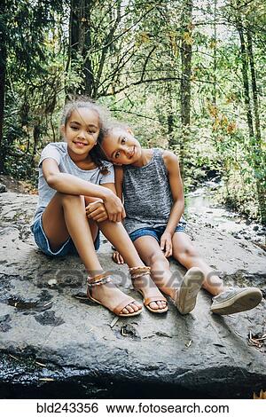 Stock Photograph - Portrait of smiling Mixed Race girls sitting on rock. Fotosearch