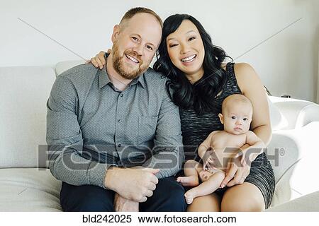 Stock Photography - Portrait of smiling parents posing with baby daughter on sofa. Fotosearch