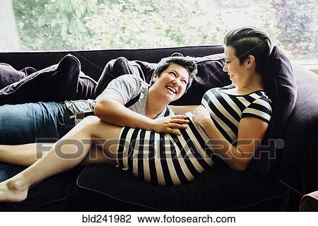Stock Image - Pregnant lesbian couple cuddling on sofa. Fotosearch