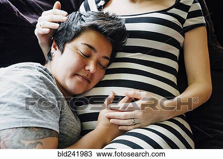 Stock Image - Pregnant lesbian couple cuddling on sofa. Fotosearch