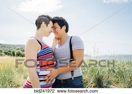 Stock Image - Pregnant lesbian couple hugging outdoors. Fotosearch