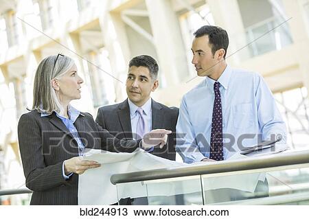 Stock Image - Serious business people with blueprint talking in lobby. Fotosearch
