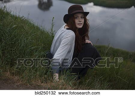 Stock Image - Serious Caucasian woman sitting on hill near lake. Fotosearch