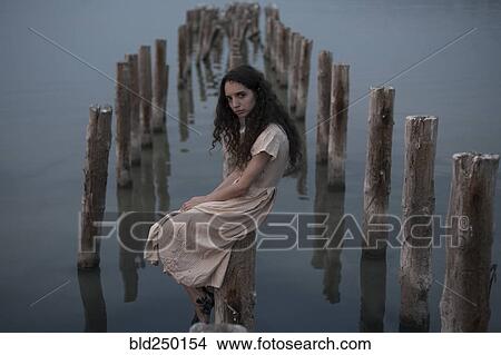 Picture - Serious Caucasian woman sitting on piling in river. Fotosearch
