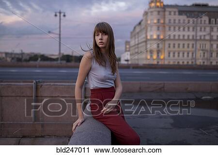 Stock Image - Serious Caucasian woman sitting on wall in city. Fotosearch