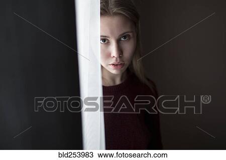 Stock Image - Serious Caucasian woman standing near curtain at window. Fotosearch