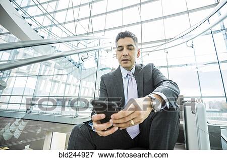 Stock Photo - Serious Mixed Race businessman sitting on staircase texting on cell phone. Fotosearch