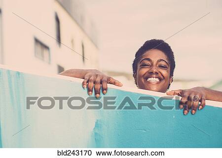 Stock Image - Smiling African American woman peering over blue wall. Fotosearch
