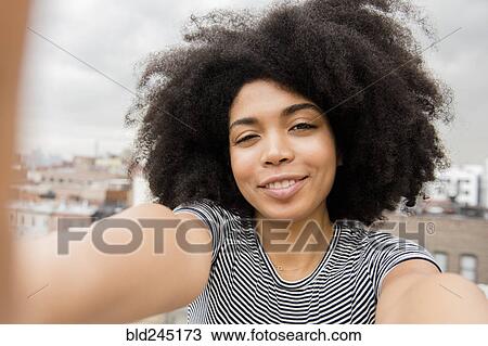 Stock Image - Smiling African American woman posing for selfie on rooftop. Fotosearch