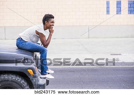 Stock Photography - Smiling African American woman sitting on hood of car. Fotosearch