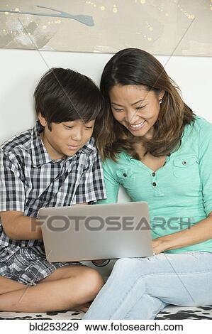 Stock Photo - Smiling Asian mother and son sitting on bench using laptop. Fotosearch