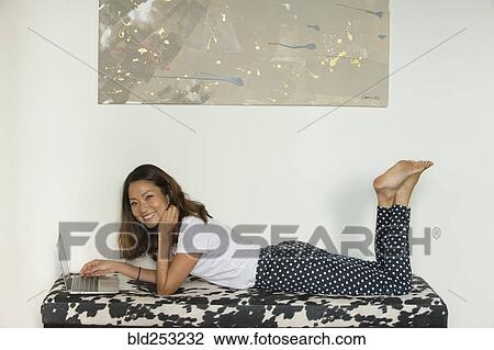 Stock Image - Smiling Asian woman laying on bench using laptop. Fotosearch