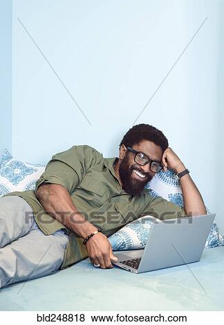Smiling black man laying on bed using laptop View Large Photo Image Stock Photo - Smiling black man laying on bed using laptop. Fotosearch
