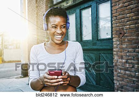 Smiling Black woman listening to cell phone with headphones in city View Large Photo Image Stock Image - Smiling Black woman listening to cell phone with headphones in city. Fotosearch