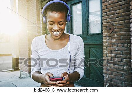 Smiling Black woman listening to cell phone with headphones in city View Large Photo Image Stock Image - Smiling Black woman listening to cell phone with headphones in city. Fotosearch