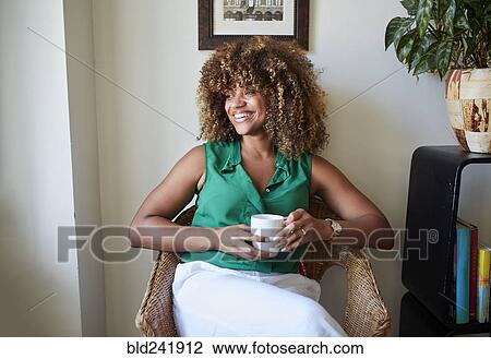 Stock Image - Smiling Black woman sitting in armchair drinking coffee. Fotosearch