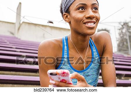 Picture - Smiling Black woman sitting on bleachers texting on cell phone. Fotosearch