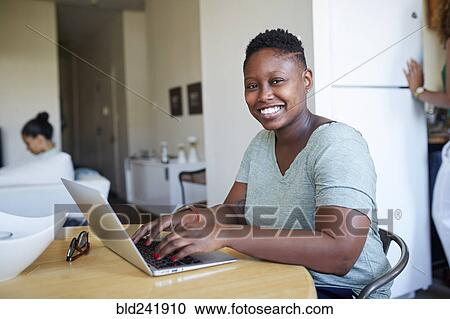 Stock Image - Smiling Black woman using laptop at table. Fotosearch