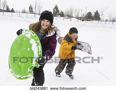 Stock Image - Smiling boy and girl carrying toboggan and snowboard in winter. Fotosearch