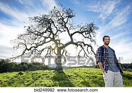 Stock Image - Smiling Caucasian man standing in field near tree. Fotosearch