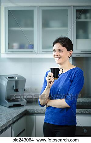 Picture - Smiling Caucasian woman drinking coffee in kitchen. Fotosearch